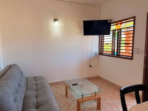 Una sala de estar con sofá, una mesa de centro de vidrio, un televisor de pantalla plana montado en la pared y una ventana con un celosía decorativa.