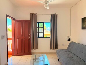 A living room with a sofa, a glass coffee table, a ceiling fan, a window with curtains, and a door. - Villas Kaab (Cozumel)