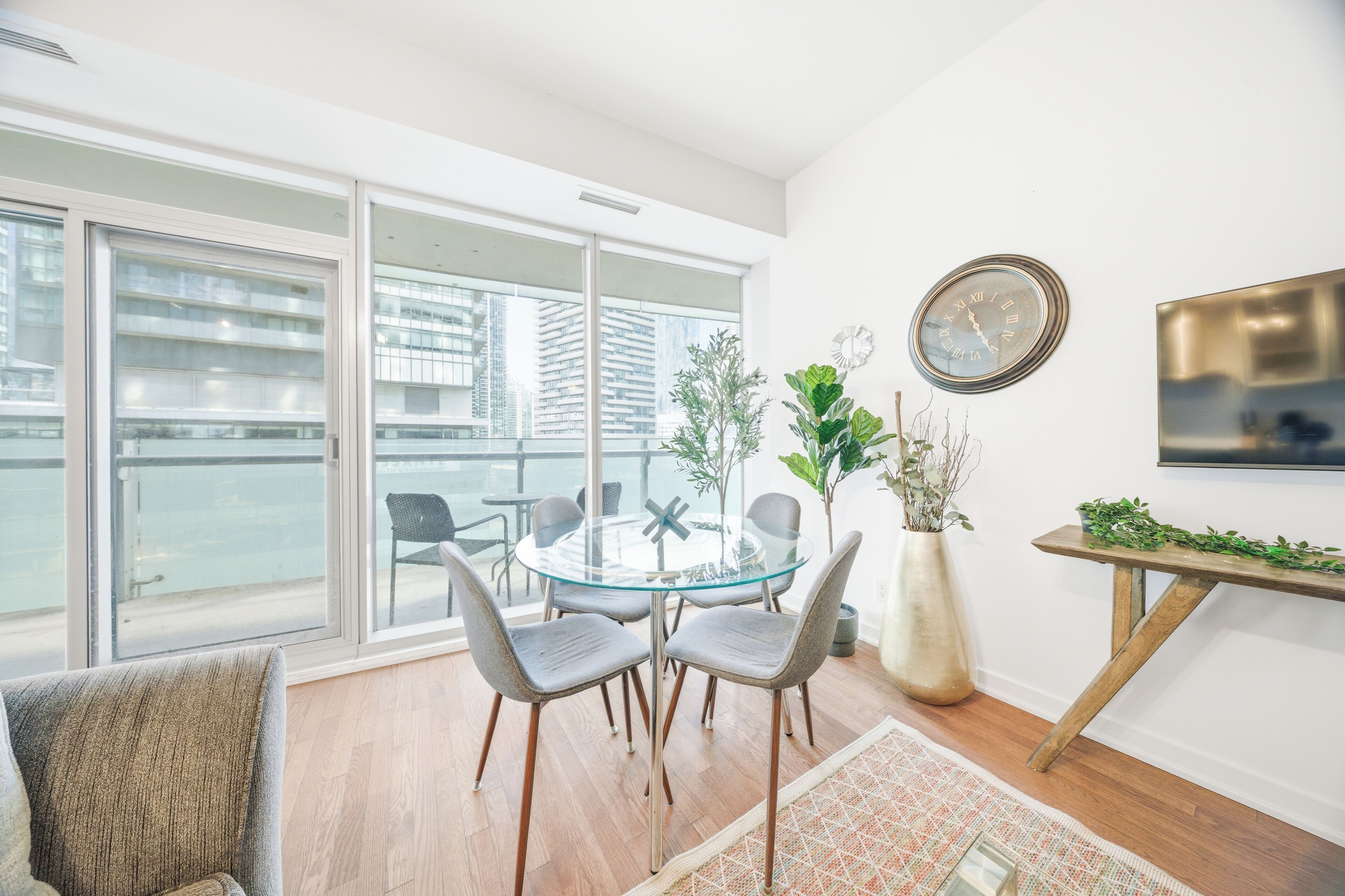Een moderne woonkamer met een glazen tafel, grijze stoelen, een groot raam en stadszicht.