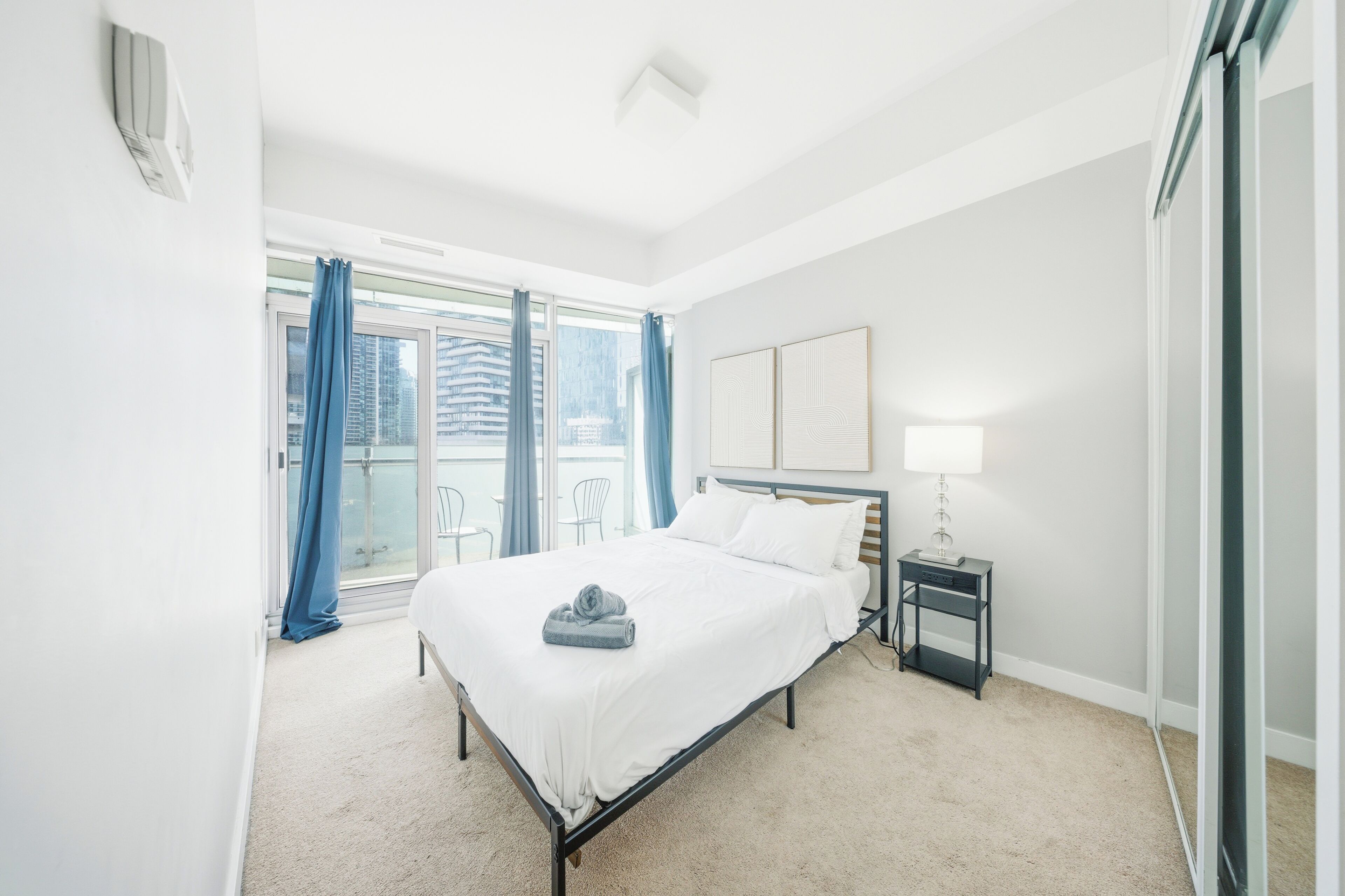 A hotel room with a bed, a nightstand, a lamp, and a view of the city through large windows.