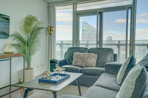 Uma sala de estar moderna com um sofá, uma mesa de centro, uma planta em vaso e vista para a paisagem urbana pelas grandes janelas.