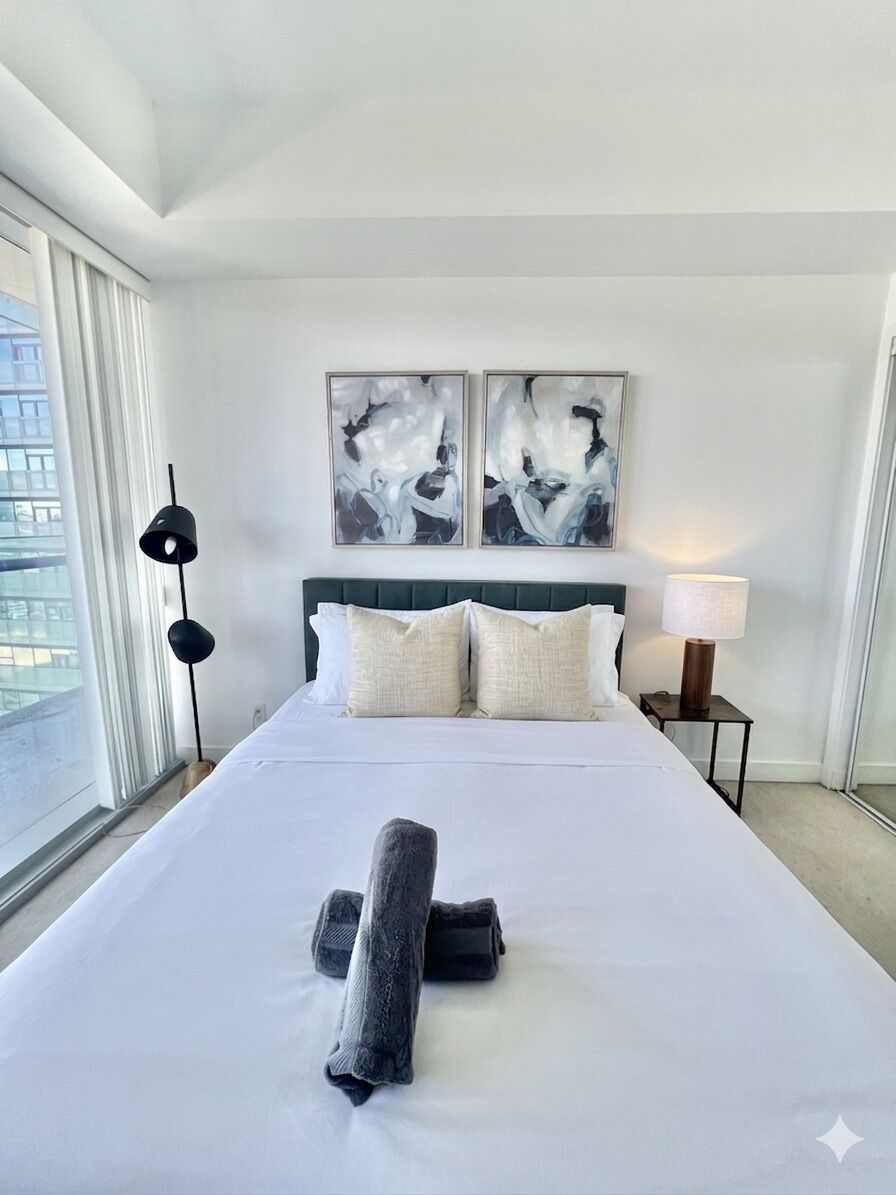 A hotel room with a large bed, two side tables, a window with curtains, and abstract artwork on the wall.