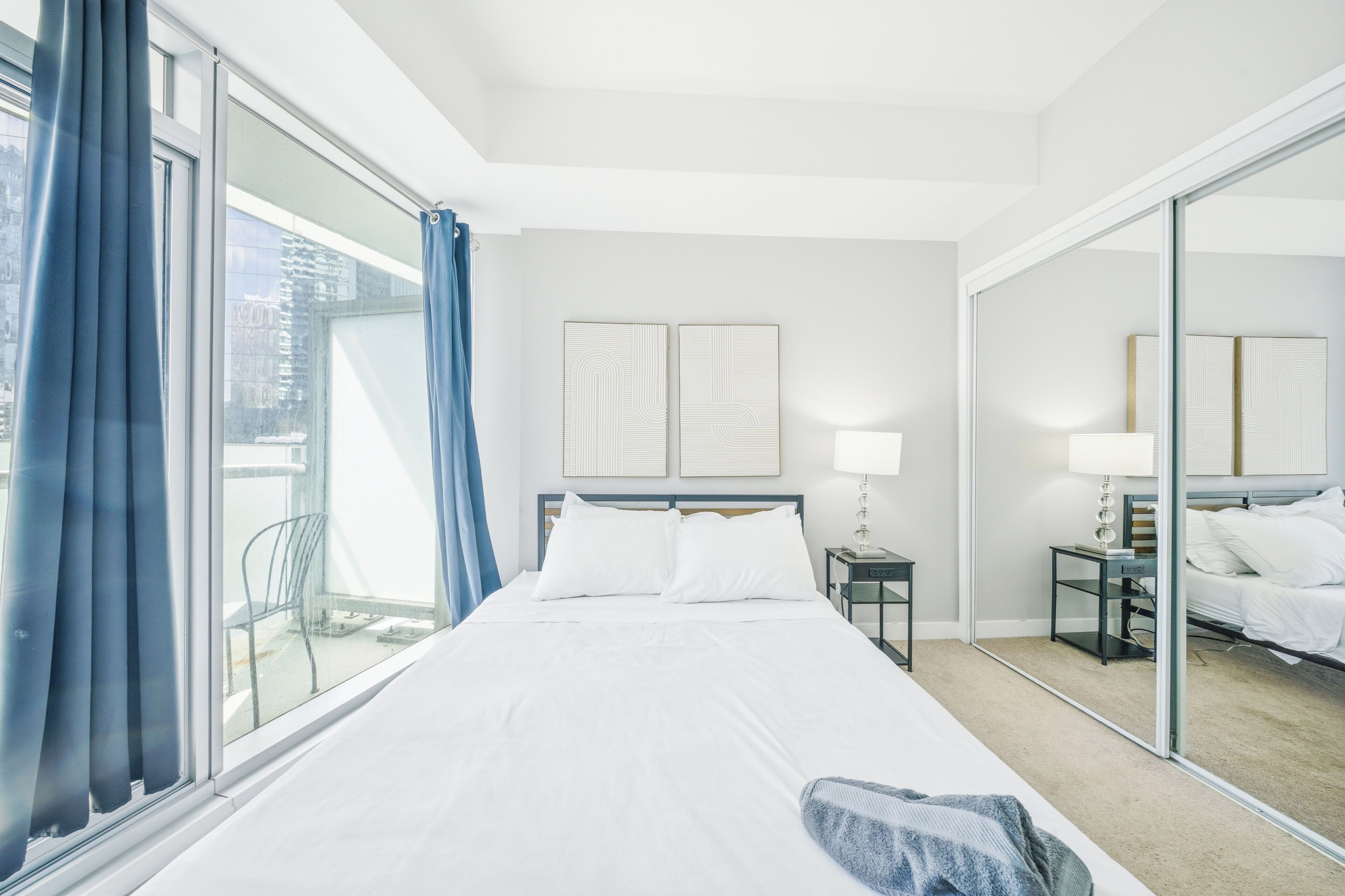 A hotel room with a large bed, a bedside table with a lamp, a mirror, and a view of the outside.