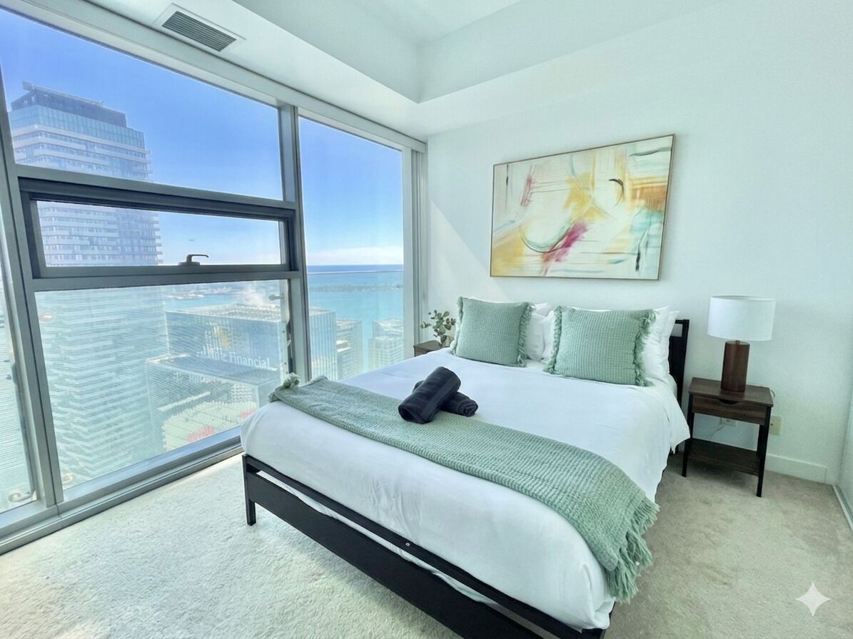 A modern bedroom with a large window, a bed with a green blanket, a nightstand, and a painting on the wall.