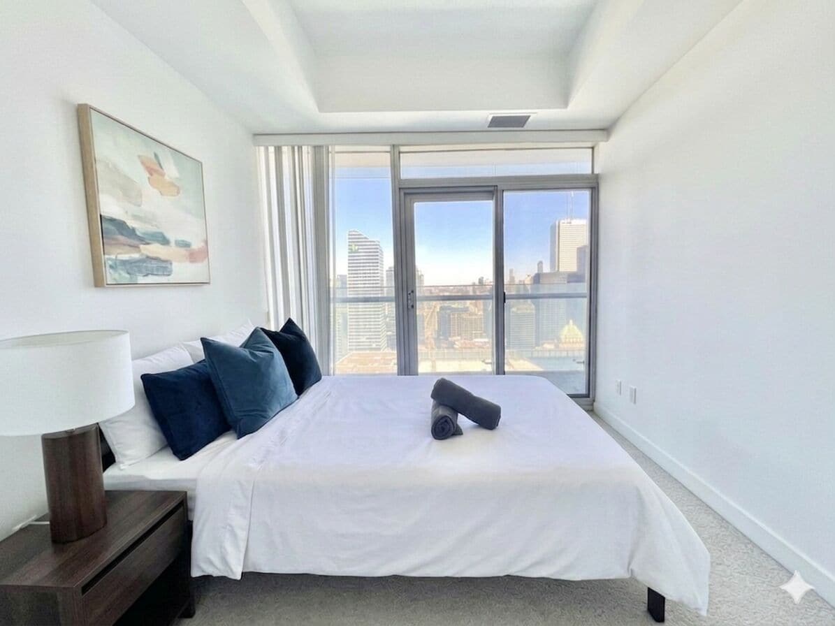 A bedroom with a bed, a nightstand, a lamp, a window with a city view, and a painting on the wall.