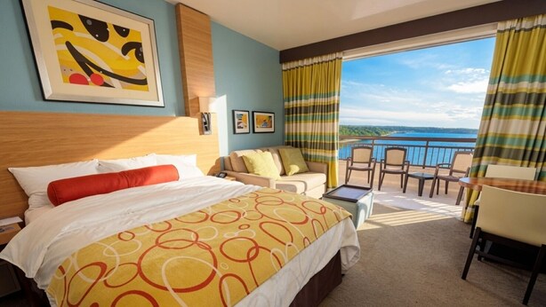 A hotel room with a bed, a sofa, a small table, and a balcony with chairs.