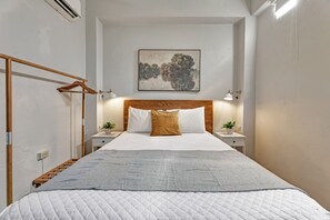 A hotel room with a bed, a wooden headboard, two bedside tables with lamps, a wall-mounted air conditioner, and a painting above the bed. - San Cristóbal View | 2BR in Old San Juan (San Juan)