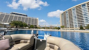 Ein Boot mit Sitzgelegenheiten und Blick auf einen Pool sowie ein Hotel mit mehreren Stockwerken und einer Brücke im Hintergrund.