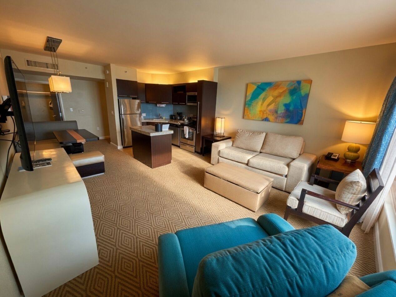 Ein Hotelzimmer mit einer Küchenzeile, einem Sofa, einem Esstisch und einem großen Fenster mit Vorhängen.