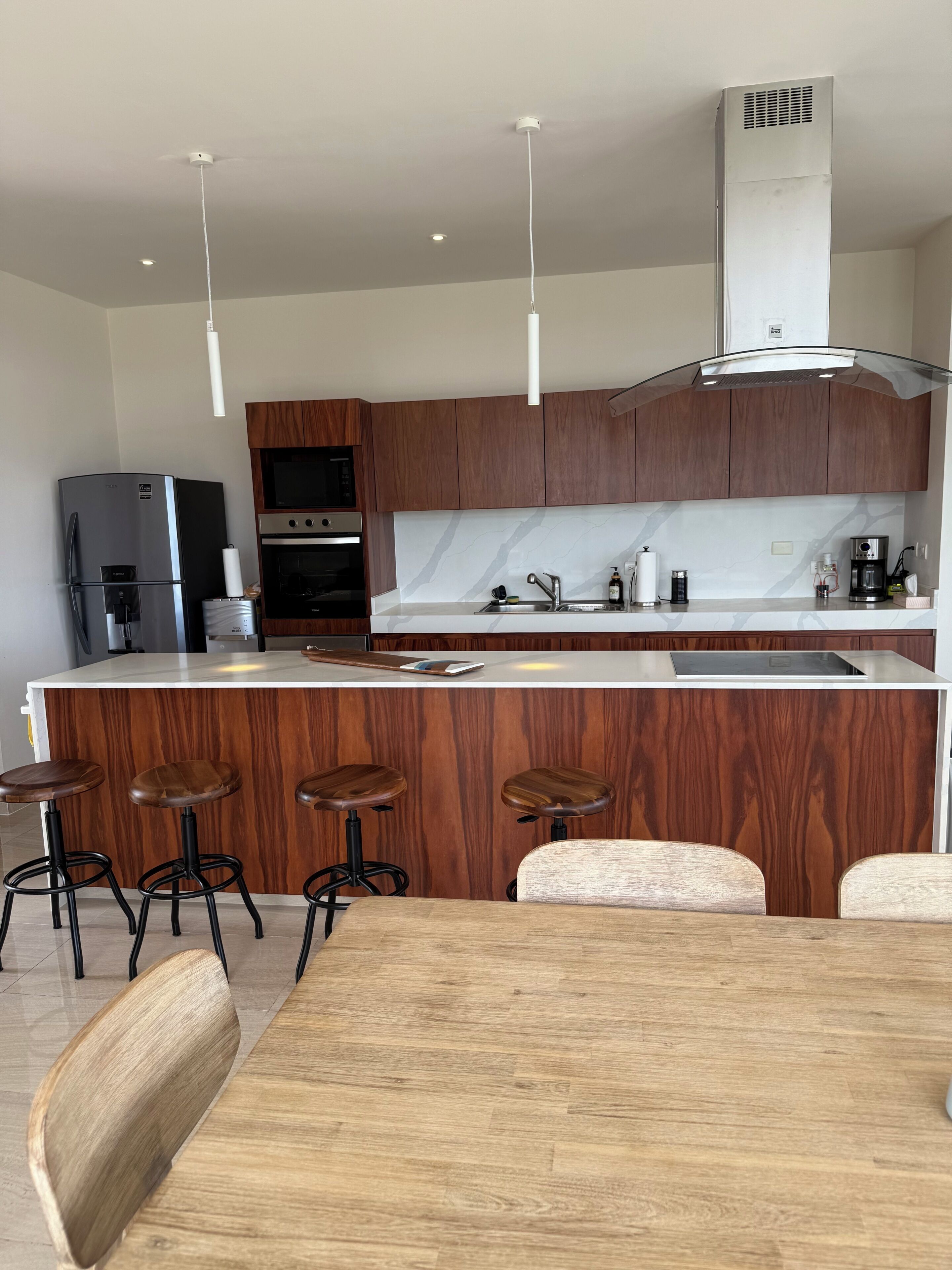 A modern kitchen with wooden cabinets, a stainless steel refrigerator, a built-in oven, a coffee maker, and a bar area with stools.