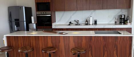 A modern kitchen with wooden cabinets, a stainless steel refrigerator, a built-in oven, a coffee maker, and a bar area with stools.
