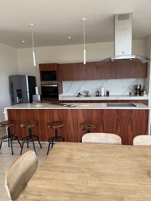 A modern kitchen with wooden cabinets, a stainless steel refrigerator, a built-in oven, a coffee maker, and a bar area with stools. - Galia Cozumel  (Cozumel)