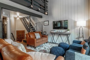 A modern living room with a brown leather sofa, blue armchairs, a staircase, a flat-screen TV, and a wooden coffee table. - Spacious 2BR + Loft Mammoth Escape 680 (Mammoth Lakes)