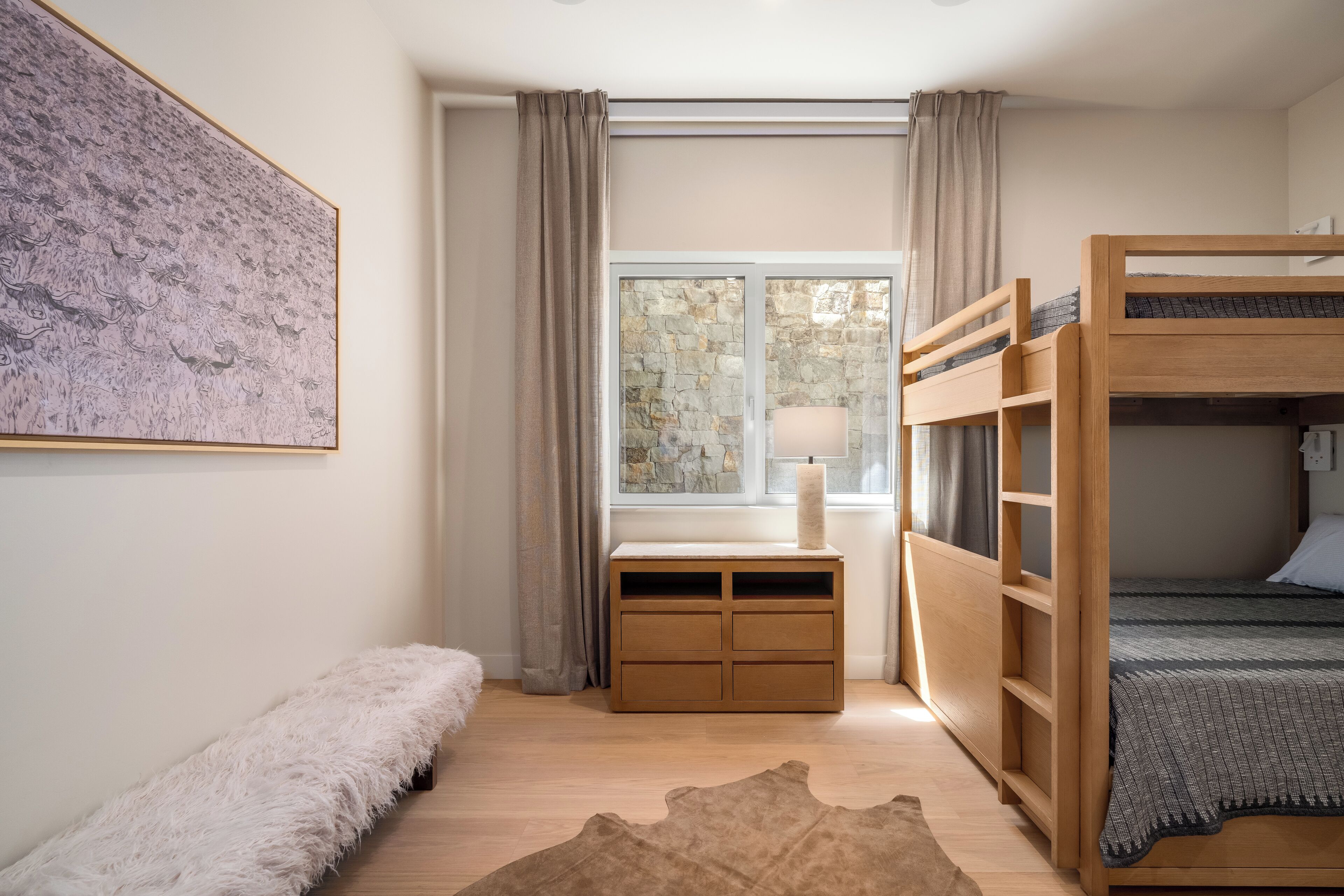 A bedroom with a bunk bed, a desk with a lamp, a fluffy bench, and a stone wall.