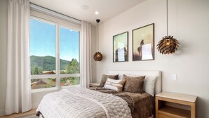 Une chambre avec une grande fenêtre, un lit recouvert d’une couette blanche, deux lampes fixées au mur et deux tableaux encadrés.