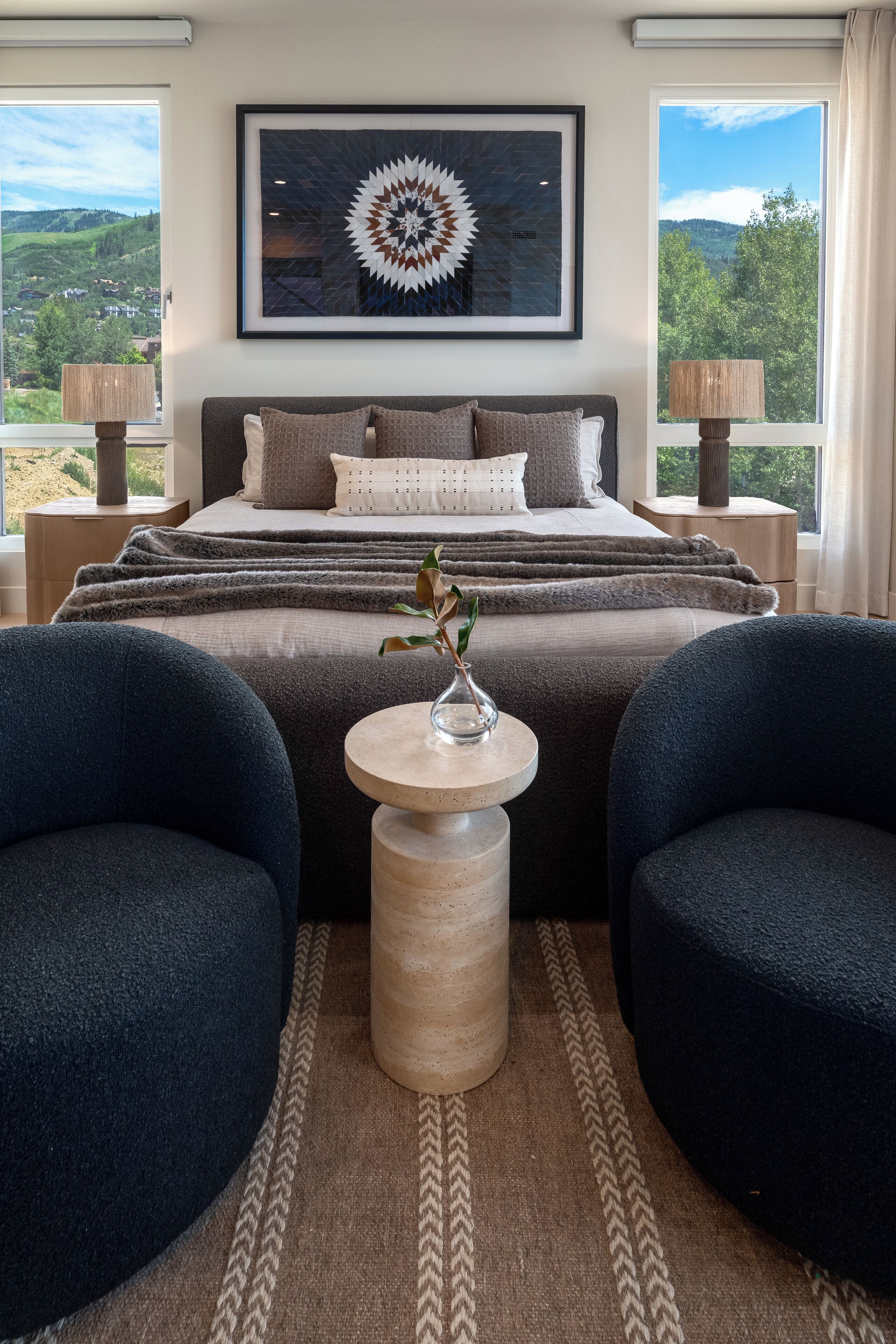 A bedroom with a large bed, two side tables, a framed artwork above the bed, and a view of the outdoors through the windows.