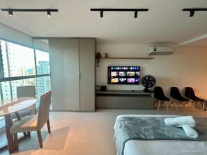 A modern hotel room with a bed, a desk, a chair, a TV, and a view of the city. - Studio de luxo no Praia de Gameleira (Recife)