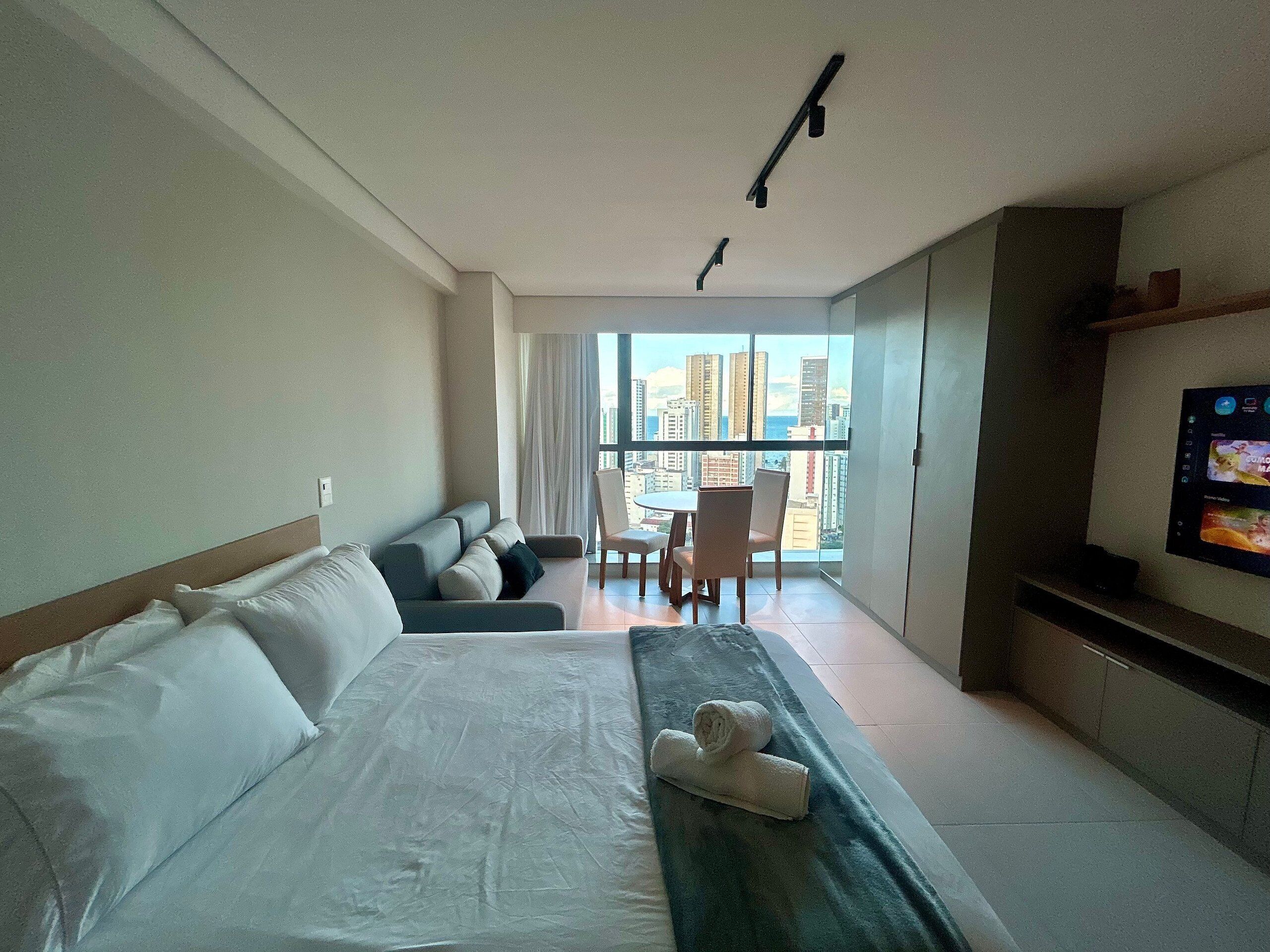 A hotel room with a bed, a sofa, a TV, a balcony with a city view, and a dining area.