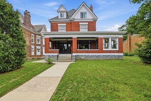 A two-story brick house with a front porch, a visible garage, and a well-maintained lawn. - Historic 1910 Pittsburgh Victorian  (Pittsburgh)