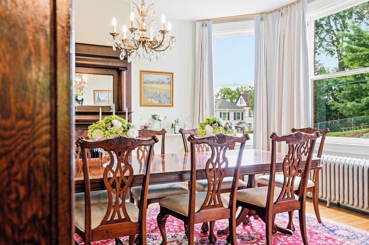 A dining room with a large table, ornate chairs, a chandelier, and a view of a house through a window.
