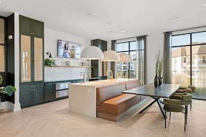 A modern kitchen with a central island, a dining area with a bench, and large windows offering a view of the surroundings. - Chic, New Apt | Pool | Long Stays (Athens)