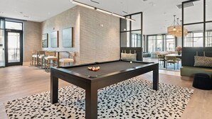 Uma mesa de bilhar em um espaço moderno com parede de tijolos, janelas amplas e um espaço de estar com sofá e poltronas.