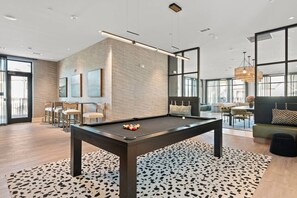 A pool table in a modern room with a brick wall, large windows, and a seating area with a sofa and chairs. - Chic, New Apt | Pool | Long Stays (Athens)