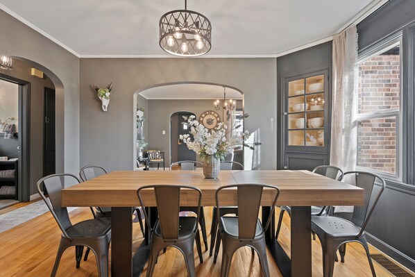 A dining area with a wooden table, metal chairs, a chandelier, and a large window with a view of a brick building.