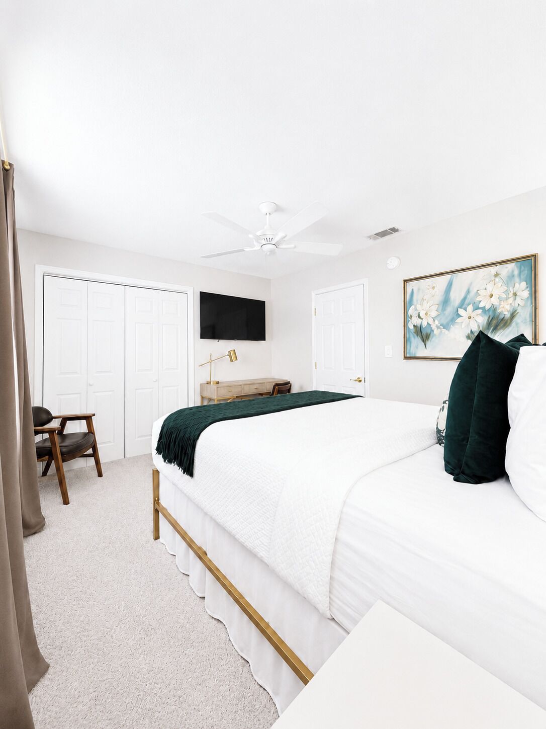 A bedroom with a bed, a desk, a chair, a ceiling fan, and a painting on the wall.