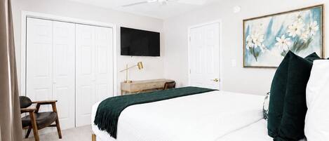A bedroom with a bed, a desk, a chair, a ceiling fan, and a painting on the wall.