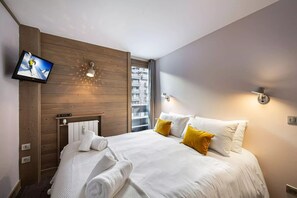 A hotel room with a bed, a television mounted on the wall, a window with a view of buildings, and a wooden wall panel. - Solaise 202–302 – Stunning Renovated Duplex in the Heart of Val d’Isère (Val-d'Isère)