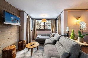 Ein modernes Wohnzimmer mit grauem Sofa, einem Holztisch, einem an der Wand befestigten Fernseher, einem kleinen Topfpflanzen und Blick auf ein Gebäude durch das Fenster.