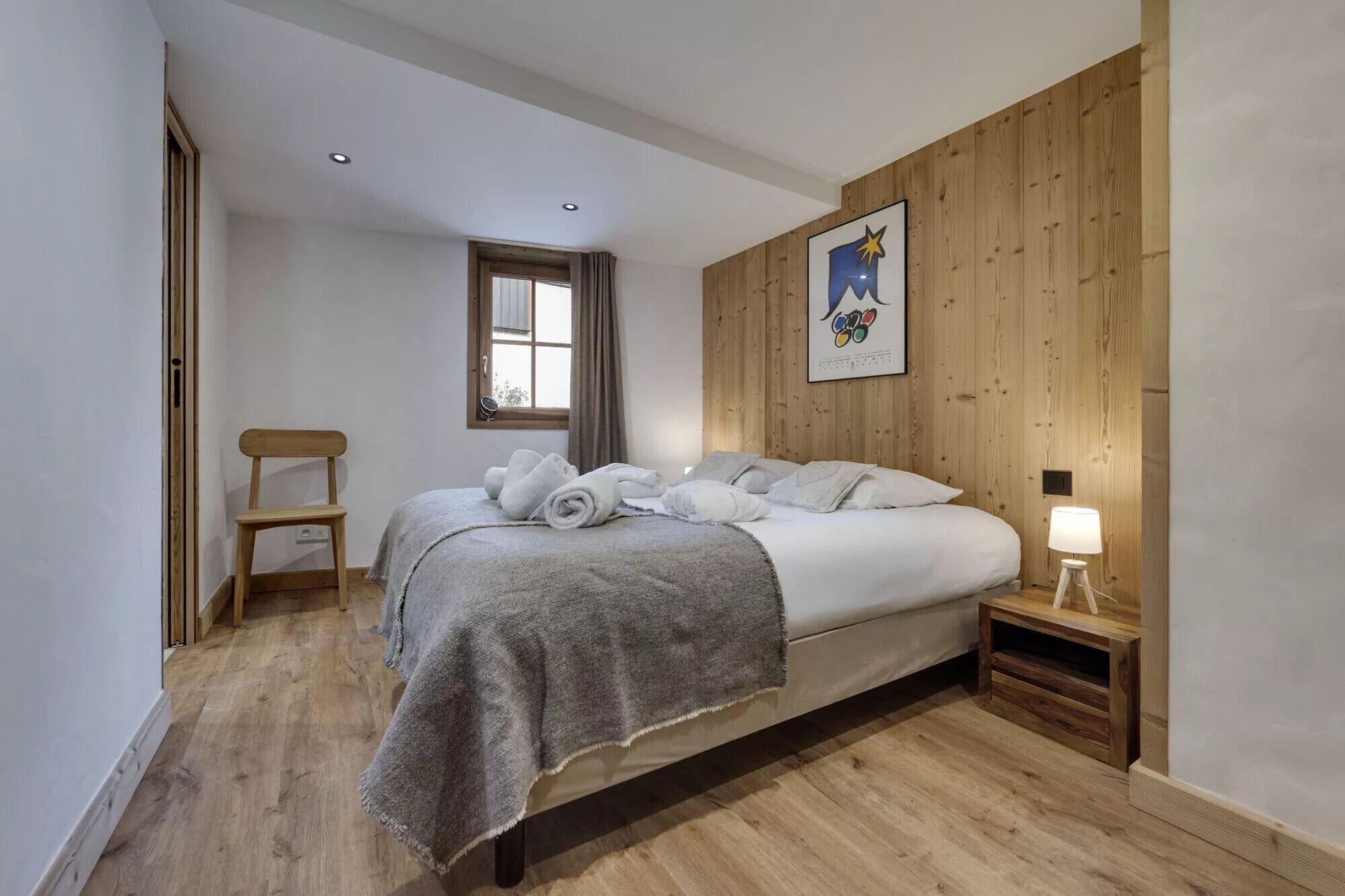 A bedroom with a wooden headboard, a bed with white linens, a bedside table with a lamp, and a window with curtains.