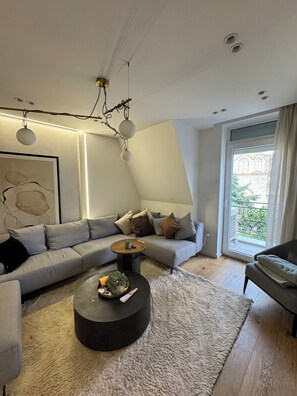 A modern living room with a sectional sofa, a round coffee table, and a large window with a view. - 2BR Skopje Apartment with Cathedral View, In The Main Square (Skopje)