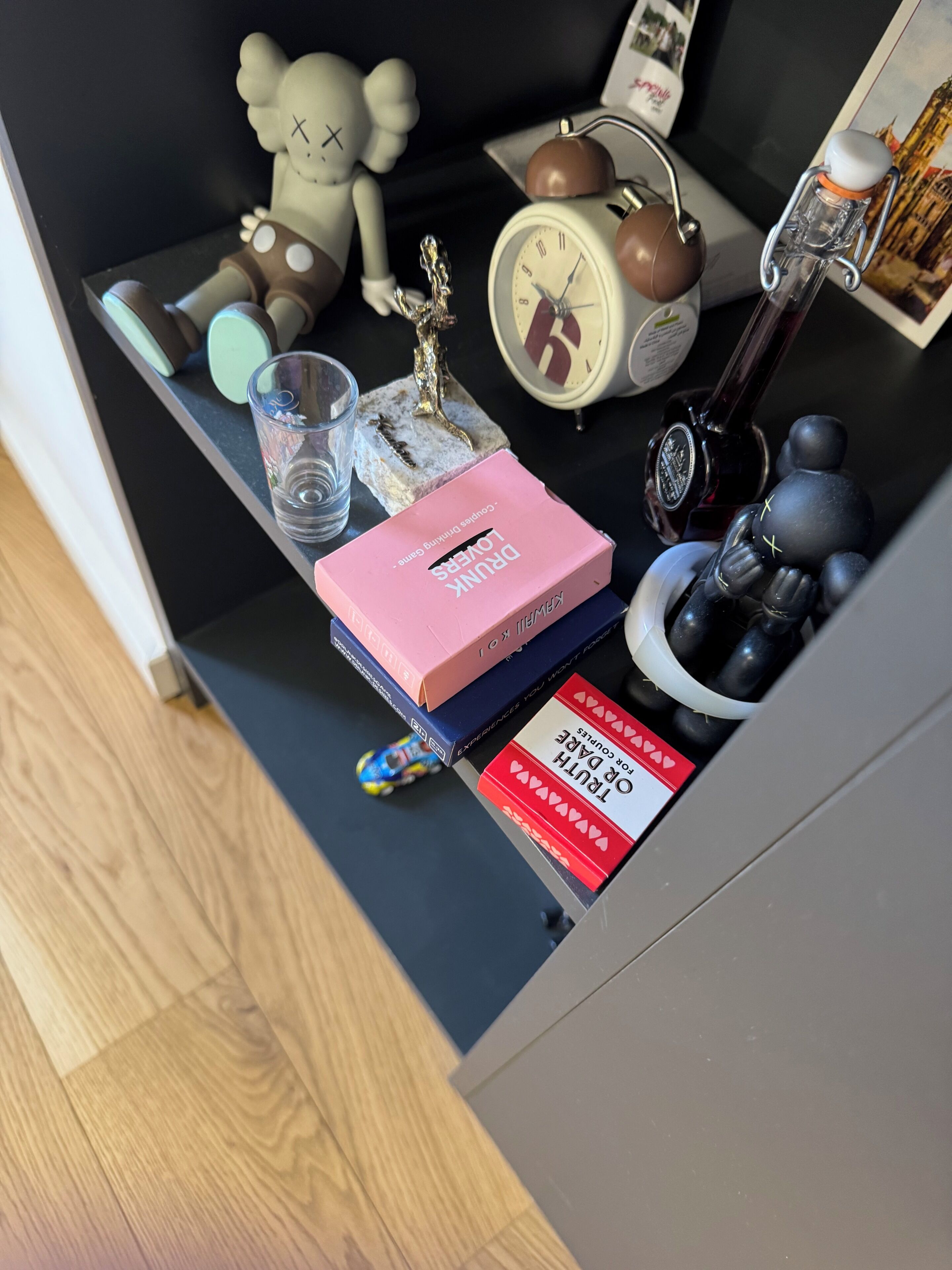 Ett litet bord med olika föremål, bland annat en rosa låda med texten ”BERUSADE ÄLSKARE”, en liten klocka, en liten statyett, ett glas och en leksaksbil.