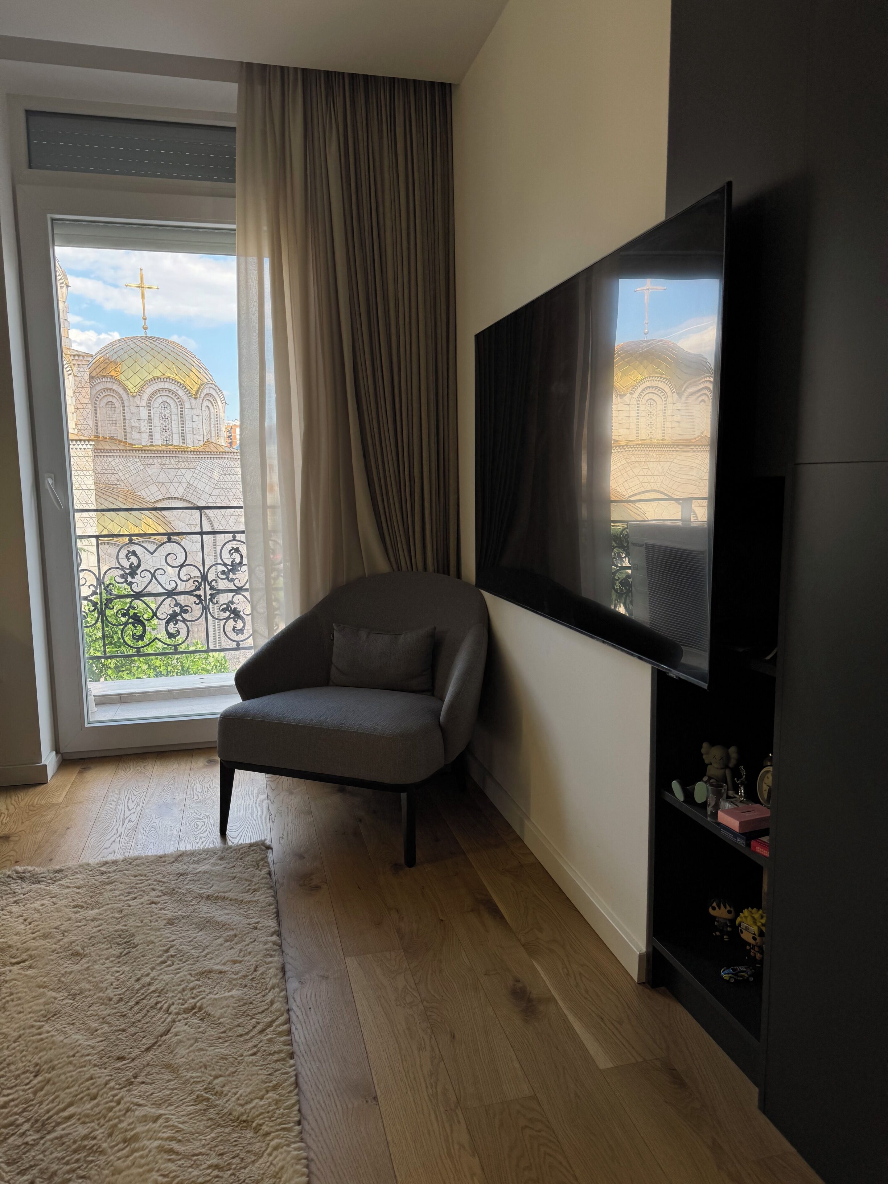 A modern living room with a grey sofa, a flat-screen TV mounted on the wall, a large window with a view of a building with a dome, and a wooden floor.