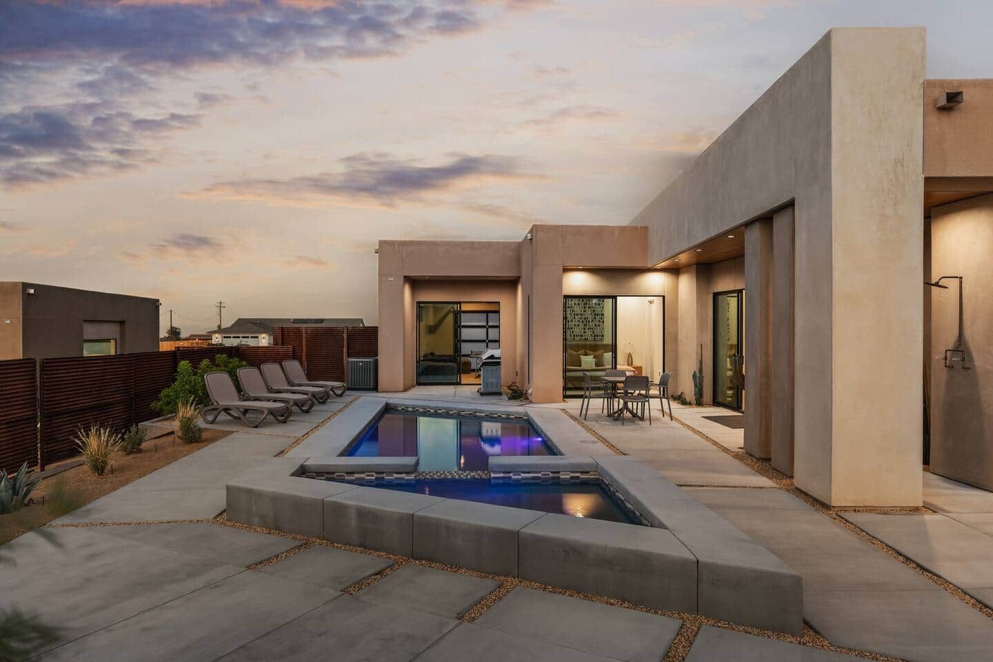 A modern outdoor pool area with lounge chairs and a dining table, surrounded by a contemporary building with large windows and a flat roof.