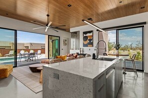 A modern kitchen with a large island, a dining area with a pool view, and a living space with a sofa and chairs. - Nightfall | Custom pool, spa, sauna, & game room (Twentynine Palms)