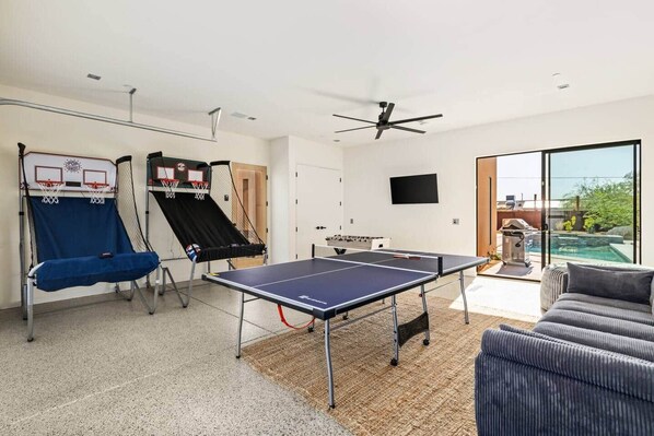 A room with a ping pong table, a basketball hoop, and a foosball table.