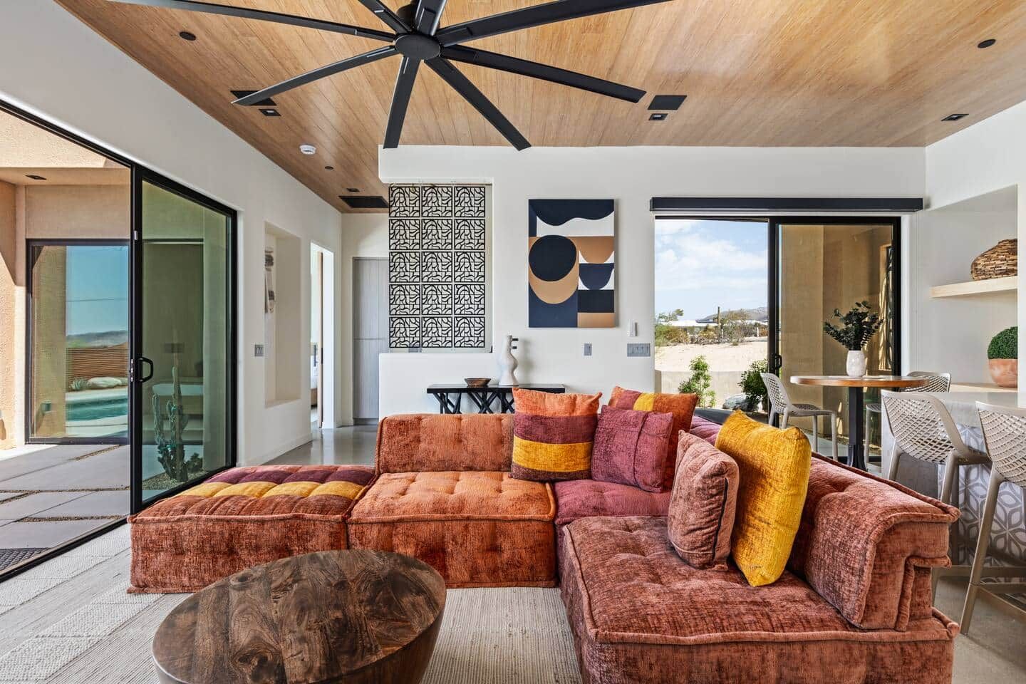 Una sala moderna con un sofá modular, un ventilador de techo de madera y un amplio ventanal con vistas al exterior.