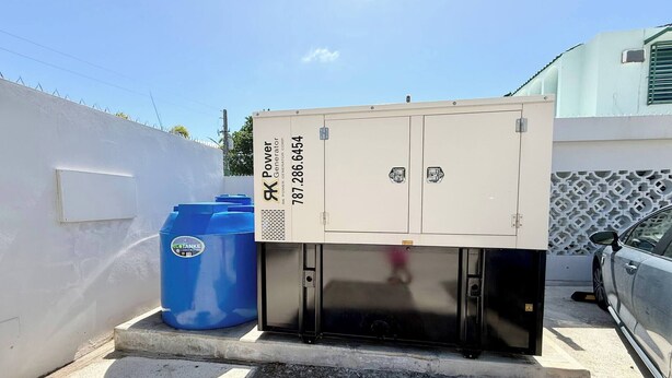 A portable power generator unit with a contact number, placed next to a blue container and a car in a parking area. - Brisamare Apt 5 | 2BR | Pool, BBQ, Generator (San Juan)