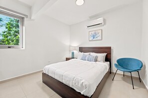Ein Schlafzimmer mit einem Bett, einem Sessel, einem Nachttisch, einer Lampe, einer Klimaanlage und einem Fenster mit Blick auf Bäume.