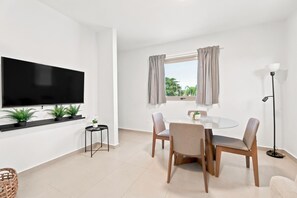 A modern living room with a flat-screen TV, a round dining table with chairs, a small side table, a floor lamp, and a window with curtains. - Brisamare Apt 4 | 1BR | Pool, BBQ, Generator (San Juan)