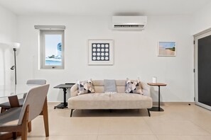A modern living room with a sofa, a dining table, and chairs. There is a window with a view of buildings, an air conditioning unit, and framed artwork on the wall. - Brisamare Apt 4 | 1BR | Pool, BBQ, Generator (San Juan)