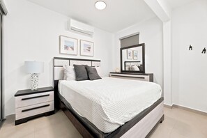 Ein Schlafzimmer mit einem Bett, Nachttischen, einer Lampe, gerahmten Bildern, einer Klimaanlage und einem Spiegel.