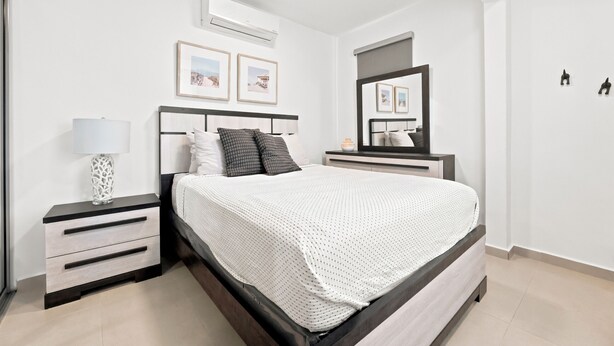 A bedroom with a bed, bedside tables, a lamp, framed pictures, an air conditioner, and a mirror. - Brisamare Collection | Pool | Sleeps 14 (San Juan)