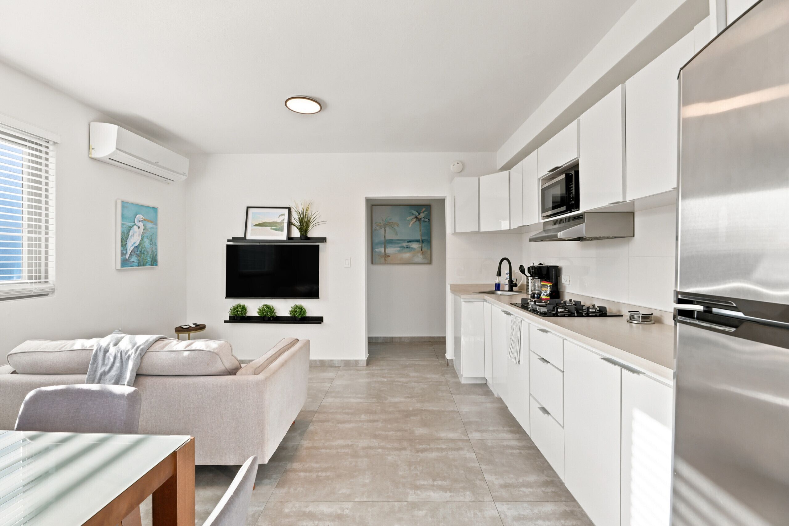 A modern kitchen with white cabinetry, a stainless steel refrigerator, a flat-screen TV mounted above the counter, and a dining area with a glass table and a sofa.