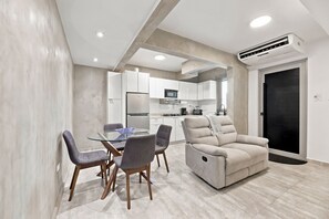 A modern living room with a glass dining table, grey upholstered chairs, a beige sofa, and a kitchen in the background. - Brisamare Apt 2 | 2BR | Pool, BBQ, Generator (San Juan)