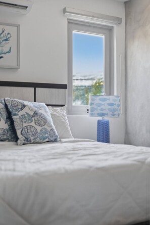 A bedroom with a bed, a window with a view of the sea, and a towel rack. - Brisamare Apt 2 | 2BR | Pool, BBQ, Generator (San Juan)