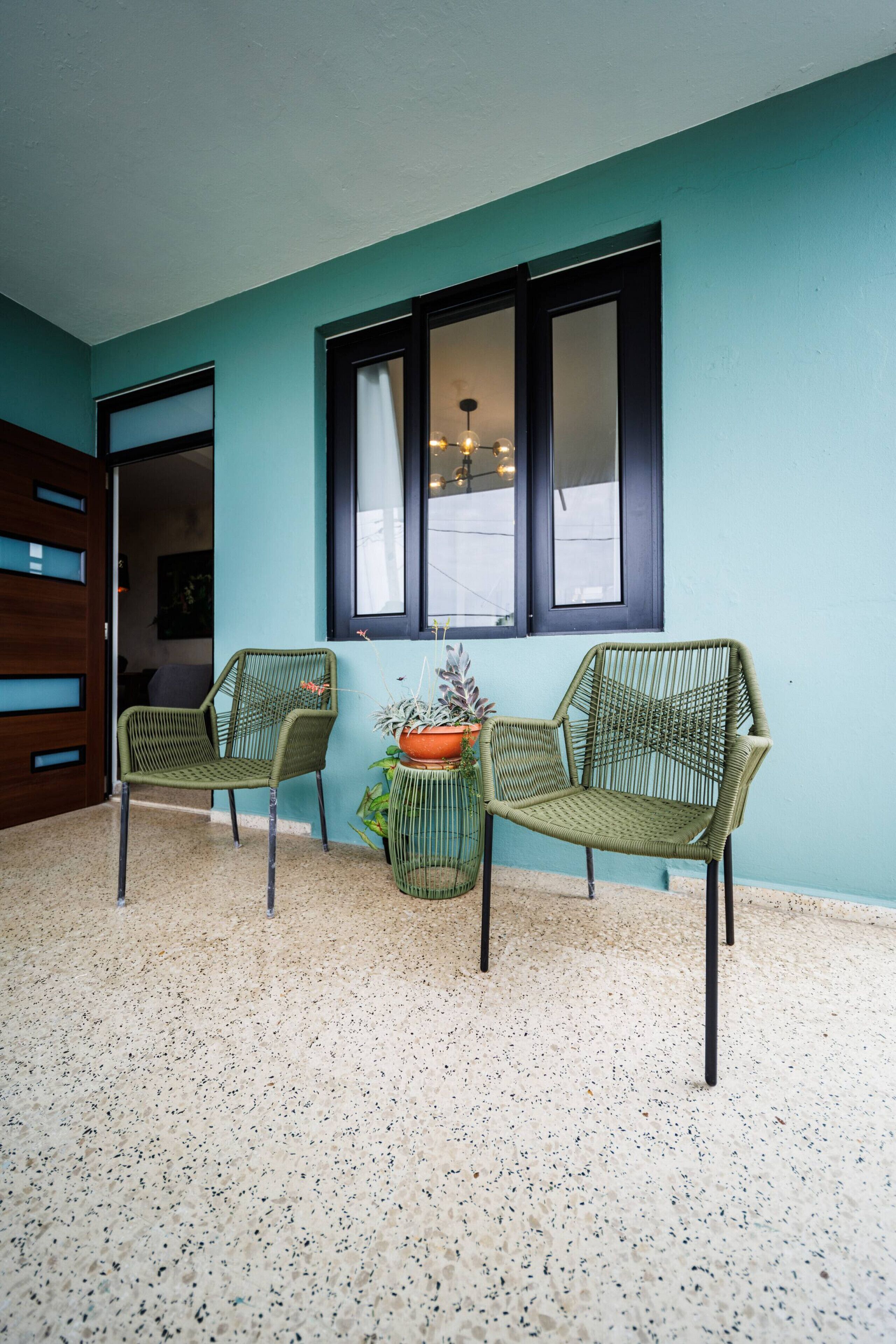 Una habitación con paredes color turquesa, dos sillas de mimbre, una mesita con una planta en maceta y una ventana con cortinas.
